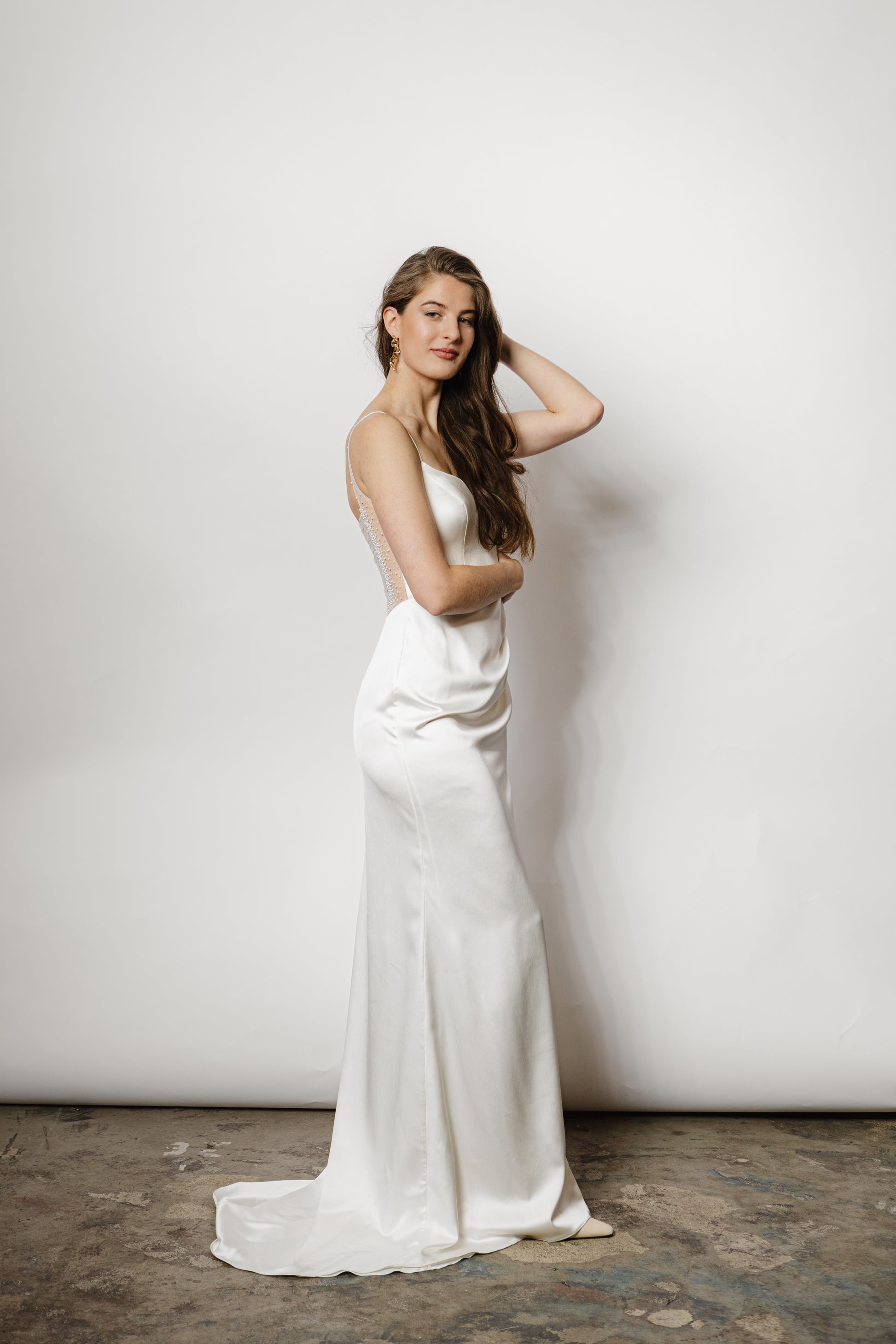 Minimalistisches Brautkleid mit Spaghettiträgern, Beinschlitz und luxuriösem Satin, der sich in einem perlig fallenden Rückendetail zeigt – moderne Eleganz für die Braut.