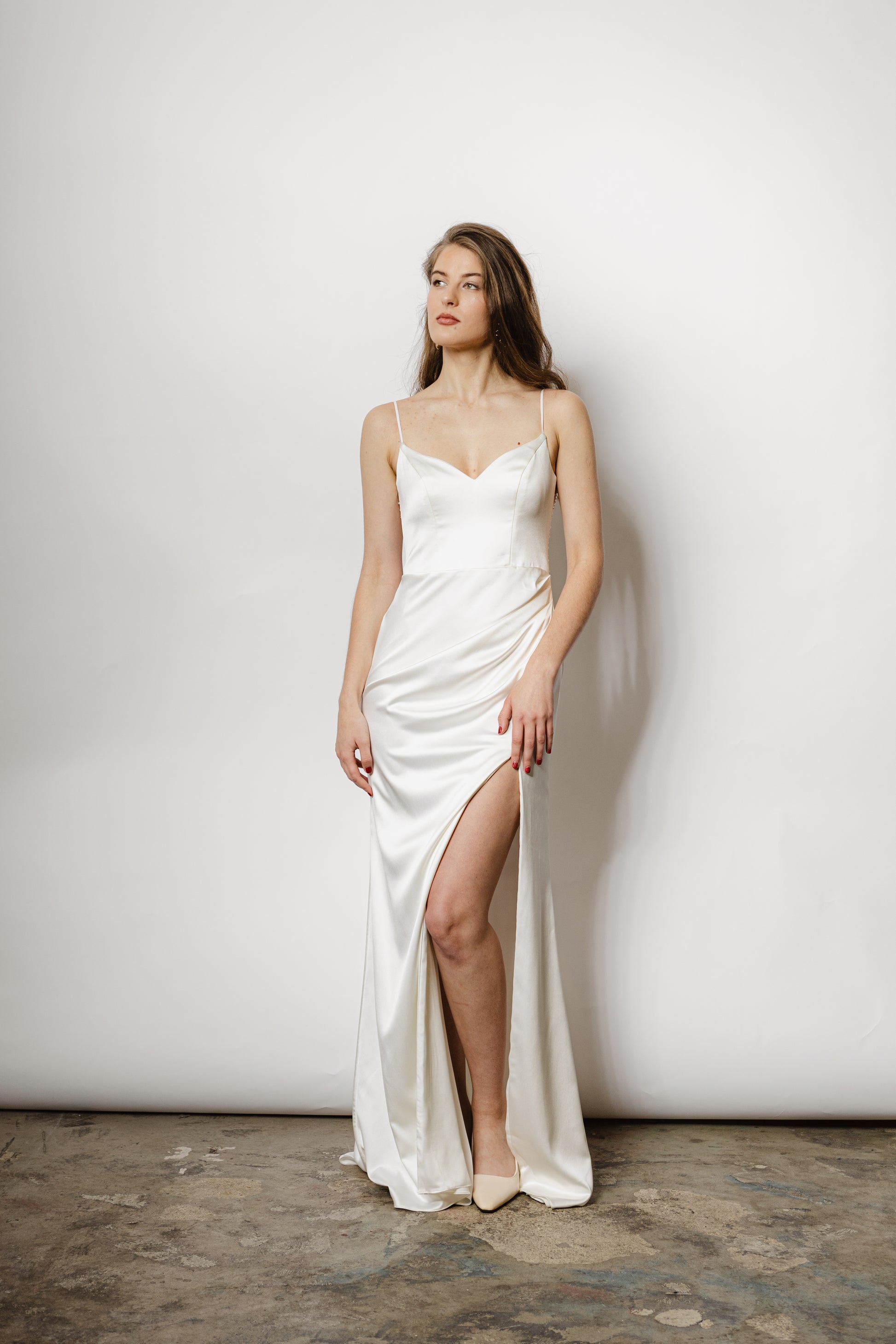 Minimalistisches Brautkleid mit Spaghettiträgern, Beinschlitz und luxuriösem Satin, der sich in einem perlig fallenden Rückendetail zeigt – moderne Eleganz für die Braut.
