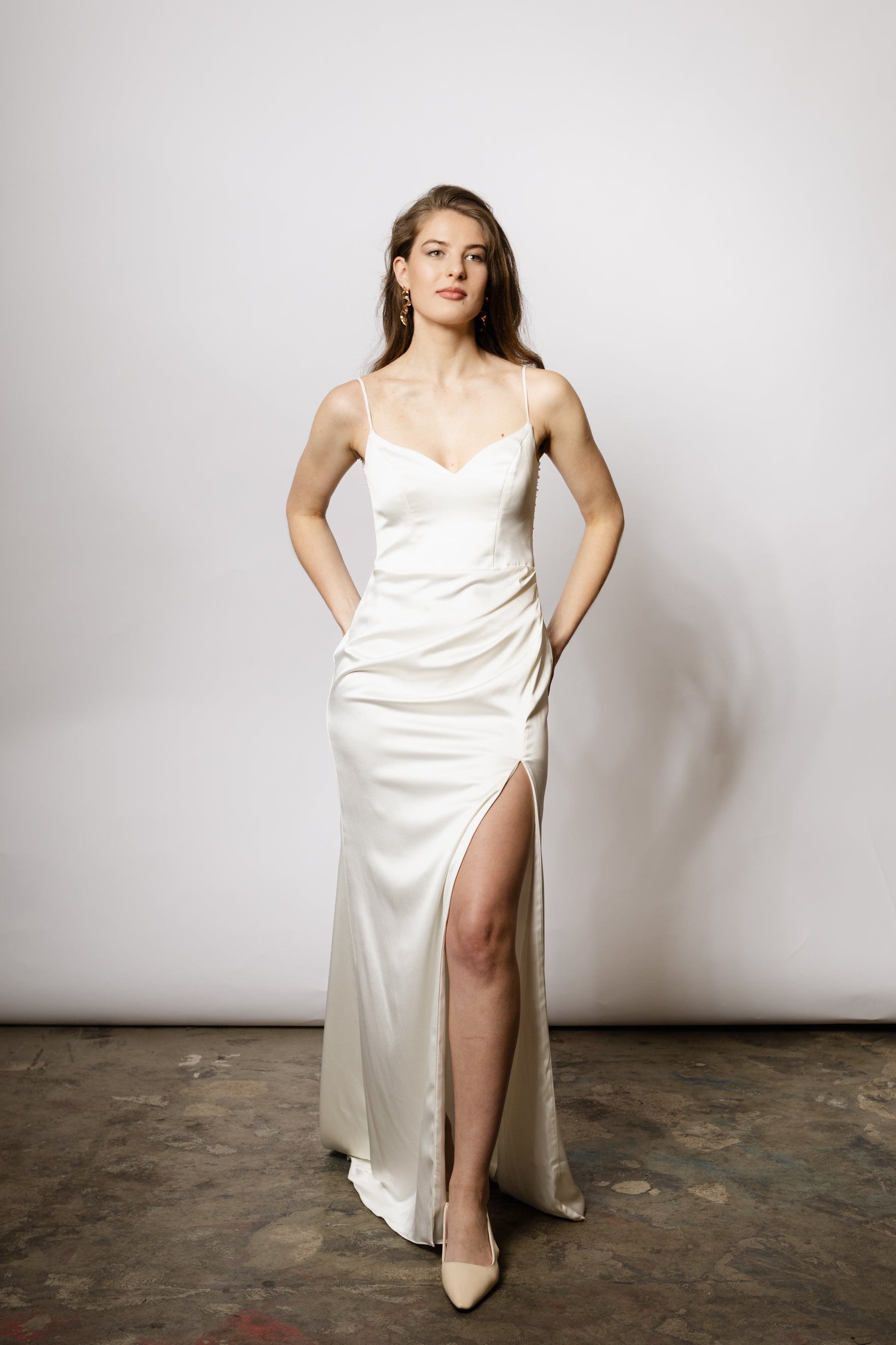 Minimalistisches Brautkleid mit Spaghettiträgern, Beinschlitz und luxuriösem Satin, der sich in einem perlig fallenden Rückendetail zeigt – moderne Eleganz für die Braut.