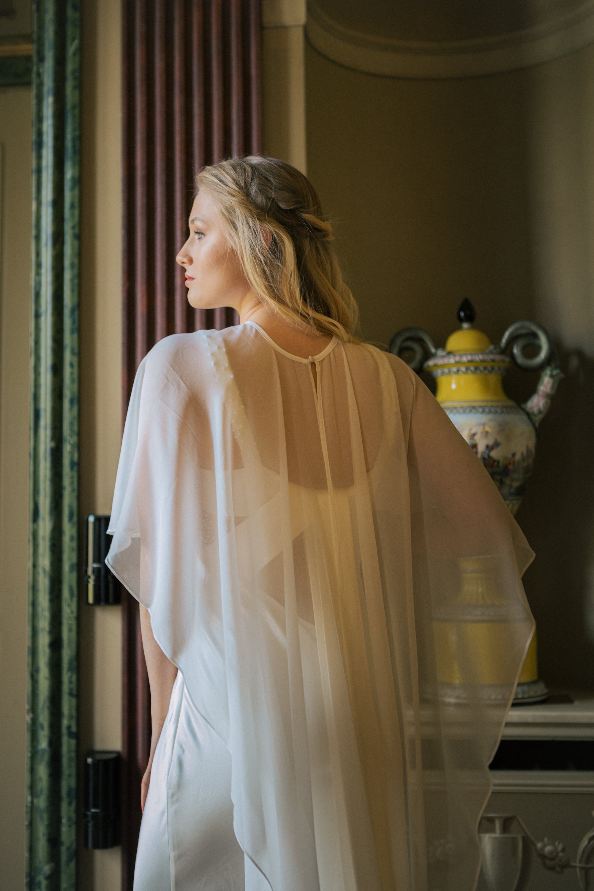 Sheer white chiffon wedding cape flowing softly over the shoulders, adding a delicate and ethereal touch to the bridal look Zarter, transparenter Chiffon-Brautcape in Weiß, der sanft über die Schultern fällt und dem Brautlook eine elegante, ätherische Note verleiht.