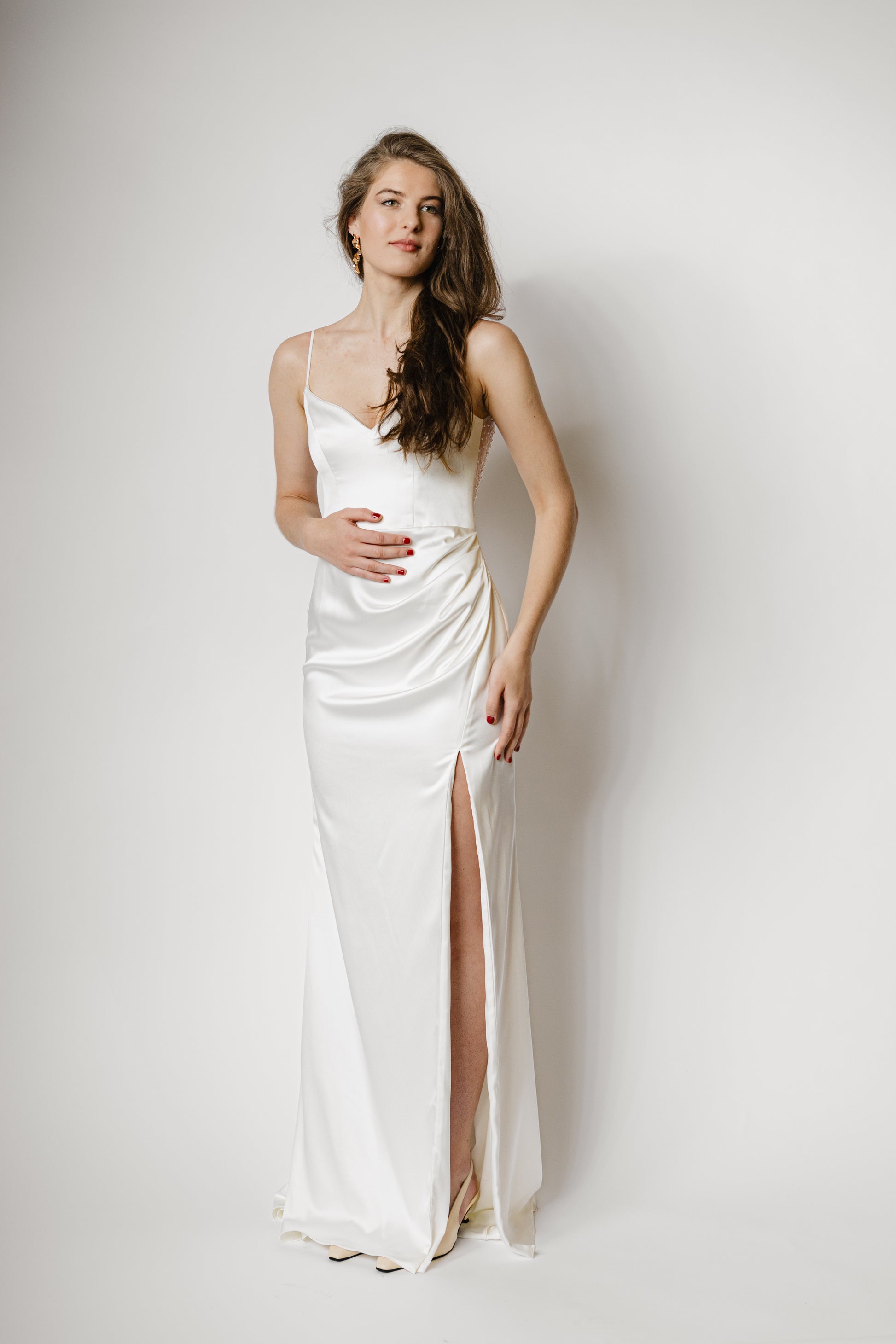 Minimalistisches Brautkleid mit Spaghettiträgern, Beinschlitz und luxuriösem Satin, der sich in einem perlig fallenden Rückendetail zeigt – moderne Eleganz für die Braut.